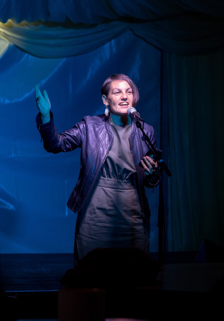 A woman standing at a microphone on a festival stage with dramatic lighting.
Photograph by Fernando Manoso.